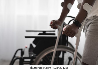 Physical Activity And Training To Recover From Injury And Illness. Adult African American Man Disabled With Crutches Walks Near Wheelchair On Large Window Background In Interior, Cropped, Close Up