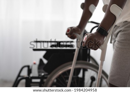 Physical activity and training to recover from injury and illness. Adult african american man disabled with crutches walks near wheelchair on large window background in interior, cropped, close up