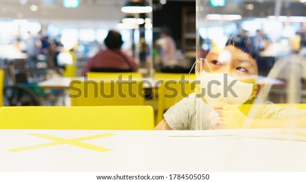 Physical distancing - New normal in Covid-19\
pandemic concept. Cute little Asian kid wear medical face mask, sit\
alone in cafeteria, Acrylic barrier, Social distance, Stop asian\
hate, Anti- AAPI.