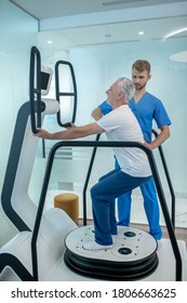 Physical Exercise, Rehabilitation. Gray-haired Man Gently Squatting Slowly Near Simulator And Young Doctor Rehabilitologist Supervising Process