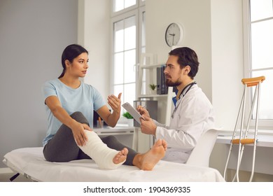 Physical injury treatment: Serious doctor listening to patient with bone fracture. Young woman with broken leg talking to traumatologist or orthopedic surgeon during medical exam in hospital or clinic - Powered by Shutterstock