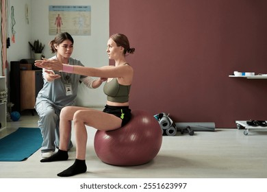 Physical therapist assisting woman with exercises on stability ball in clinical setting, focusing on achieving balance and muscle strength. Equipment visible in background - Powered by Shutterstock