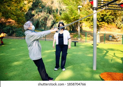 A physical therapist or professional caregiver watches closely as a senior adult woman exercises on an outdoor fitness circle. Women wear medical masks due to the pandemic. - Powered by Shutterstock