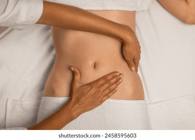 A physical therapists hands are assisting a patient by guiding and supporting their abdomen during rehabilitation exercises. The patient is lying down, wearing a white top and partially visible pants - Powered by Shutterstock