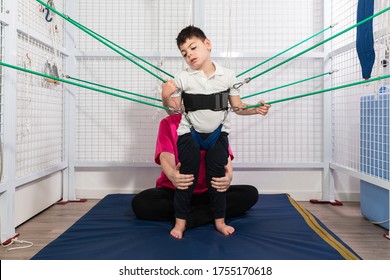 Physical Therapy Room, Physically Disabled Child Exercising With Elastic Cords, Physical Therapist Doing Rehabilitation To His Patient On Blue Mattress. Protective Mask And Gloves Coronavirus.