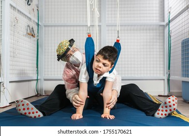 Physical Therapy Room, Physically Disabled Child Exercising With Elastic Cords, Physical Therapist Doing Rehabilitation To His Patient On Blue Mattress. Protective Mask And Gloves Coronavirus.