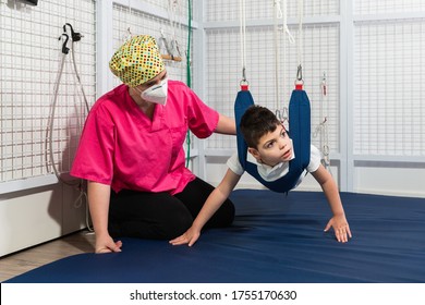 Physical Therapy Room, Physically Disabled Child Exercising With Elastic Cords, Physical Therapist Doing Rehabilitation To His Patient On Blue Mattress. Protective Mask And Gloves Coronavirus.
