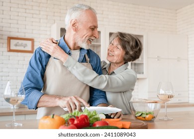 Physical touch - love language. Old elderly wife hugging embracing cuddling with husband, looking at each other helping him prepare dinner, cooking vegetable salad together - Powered by Shutterstock