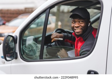 Physical working - package delivery. Positive smiling African-American man in black baseball hat sitting on driver's seat of a white van. View through vehicle window. High quality photo - Powered by Shutterstock