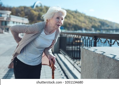 Physically Exhausted Elderly Woman Leaning On A Cane And Looking Into Vacancy While Struggling From A Physical Discomfort In A Low Back Zone.