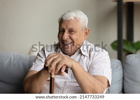 Physically optimistic grandfather hold walking stick smile at camera, looks positive, rest alone on sofa at home. Healthcare, medical insurance for older citizen, recover after trauma concept