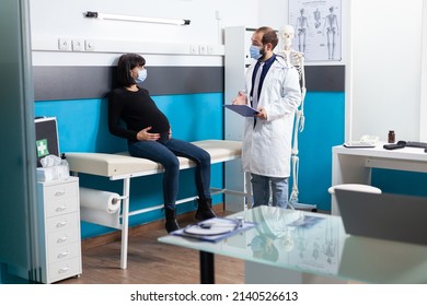 Physician With Clipboard Information Consulting Expectant Woman In Office. Patient With Pregnancy Belly And Medic Discussing Childbirth Support At Checkup Examination During Covid 19 Pandemic.