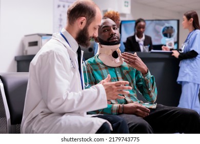 Physician Consulting Hurt Person With Neck Collar Foam, Sitting In Waiting Room At Reception Lobby. Doctor Helping Injured Man Wearing Cervical Brace For Support, Checkup In Waiting Area.