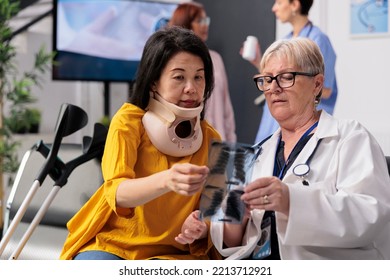 Physician Consulting Patient With Cervical Neck Collar In Hospital Waiting Room, Looking At Lungs Xray Exam, Senior Medic And Patient Discussing About Healthcare At Checkup Visit