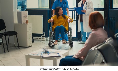 Physician Doing Medical Consultation With People In Waiting Area Lobby, Giving Support To Woman With Disability In Wheelchair. Consulting Asian Patients At Facility. Handheld Shot.