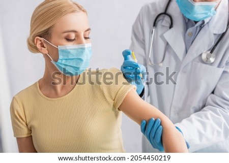 physician in latex gloves and white coat giving injection of vaccine to woman in protective mask