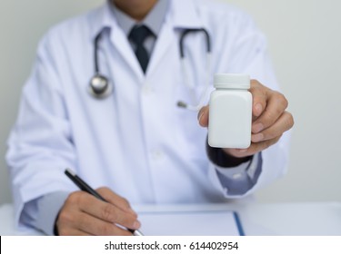 Physician Medicine Doctor Or Pharmacist Sitting At Worktable, Holding Or Showing  Bottle Of Pills In Hand And Writing Prescription On Special Form. Medical Care, Pharmacy Or Health Insurance Concept