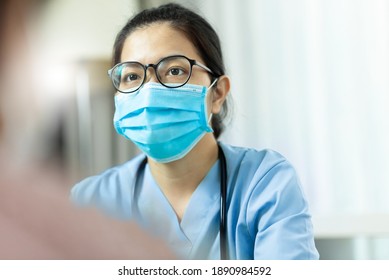 Physician Or Nurse Talking With Someone At Clinic During Pandemic Covid-19 Or Coronavirus. Asian Woman Doctor Wear Eyeglasses And Surgical Mask Giving Advice To Patient In Medical Room At Hospital.