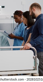 Physician Pediatric Doctor Monitoring Disease Symptoms While African American Nurse Checking IV Drip Bag In Hospital Ward. Sick Little Kid Resting In Bed Recovering After Medicine Surgery