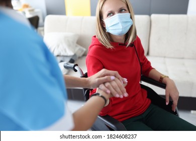 Physician Psychotherapist Holds Hand Of Disabled Young Woman In Wheelchair At Home. Working In Rehabilitation Center During Covid 19 Pandemic Concept.