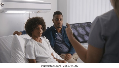 Physician reports X-Ray results to clients using tablet computer. African American teenager lies on bed and talks with doctor. Patient recovering after operation. Medical staff work in hospital ward. - Powered by Shutterstock