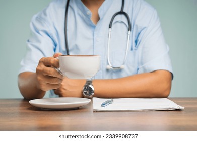 A physician with a stethoscope holding a coffee cup while sitting at the desk in the hospital. Medical, Beverage, and relaxation concepts. - Powered by Shutterstock
