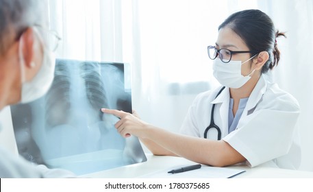 Physician Talking And Looking At X Ray Film With Senior Man At Clinic Pandemic Virus. Asian Woman Doctor Wear Eyeglasses And Surgical Mask Discussing With Elderly Patient In Medical Room At Hospital