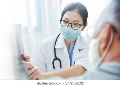 Physician Talking With Senior Man At Clinic Pandemic During Covid-19 Or Coronavirus. Asian Woman Doctor Wear Eyeglasses And Surgical Mask Giving Advice To Elderly Patient In Medical Room At Hospital
