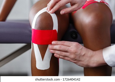 Physiotherapist Applying Kinesiology Tape To Patient Knee.Therapist Treating Young Female African American Athlete. Kinesiology Taping. Post Traumatic Rehabilitation,sport Physical Therapy.