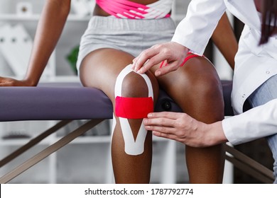 Physiotherapist Applying Kinesiology Tape To Patient Knee.Therapist Treating Young Female African American Athlete. Kinesiology Taping. Post Traumatic Rehabilitation,sport Physical Therapy.