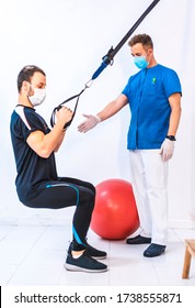 Physiotherapist In Blue Gown And A Patient In A Squat Working The Arms With Rubber Bands. Physiotherapy With Protective Measures For The Coronavirus Pandemic, COVID-19. Osteopathy, Sports Chiromassage