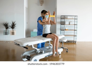 Physiotherapist Doctor In Lab Coat Assists To Exercise The Arms With Yellow Elastic Rubber To Young Woman On A Stretcher At Rehabilitation Clinic. Medical Treatments During Coronavirus Pandemic.