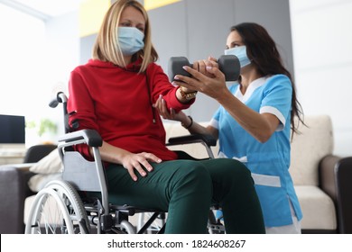 Physiotherapist Doctor In Medical Mask Helps To Raise Dumbbell To Disabled Woman In Wheelchair At Home Portrait. Rehabilitation Of Disabled People At Home During Epidemic And Coronavirus Infection