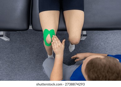 A physiotherapist explains next steps while the female patient sits on a treatment table with green knee tape. Plenty of copy space, neutral tones, and a personalized rehab care feeling.