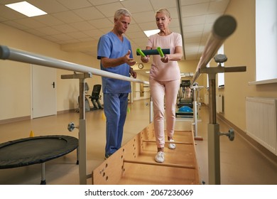 Physiotherapist Helping Mature Patient Walk Between Parallel Bars In Hospital