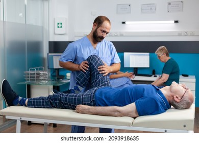 Physiotherapist Helping Old Man With Leg Injury To Recover At Kinetotherapy Clinic. Chiropractor Giving Support To Retired Patient With Knee Pain And Muscle Pressure For Recovery.