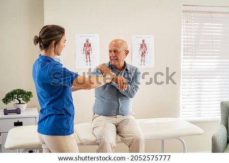 Physiotherapist helping senior man with elbow exercise in clinic. Chiropractic checking elbow and shoulder joint pain of old patient. Elderly man undergoing physiotherapy treatment for shoulder injury