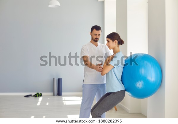 Physiotherapist helping young woman do wall squats\
with fit ball to get rid of backache and regain spinal health.\
Young female patient doing back exercise using fitball in physio\
room of modern\
clinic