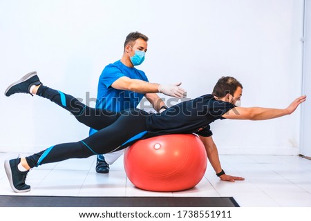 Physiotherapist with mask and a patient stretching on a ball. Physiotherapy with protective measures for the Coronavirus pandemic, COVID-19. Osteopathy, sports chiromassage