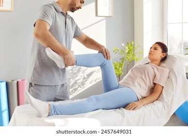 Physiotherapist treats leg injury and helps patient perform exercises for lower limbs. Female patient lies on medical couch in rehabilitation room and male doctor holds her foot and stretches her leg. - Powered by Shutterstock