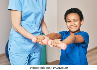 Physiotherapist Working With Child Patient In A Rehab Clinic. Boy Raises Dumbbells, Strengthening Arm Muscles And Developing Joints