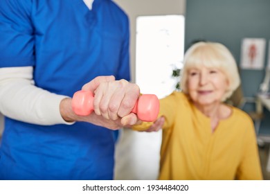 Physiotherapist Working With Female Patient In A Rehab Clinic. Elderly Woman Raises Dumbbells, Strengthening Arm Muscles And Developing Joints. Dumbbell In Focus