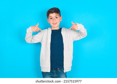 Pick me! Confident, self-assured and charismatic caucasian kid boy wearing grey hoodie over blue background promoting oneself as wanting role smiling broadly and pointing at body.