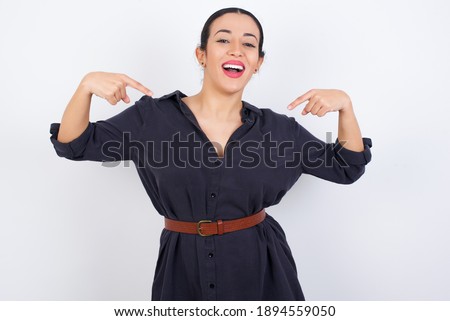 Pick me! Confident, self-assured and charismatic young beautiful Arab woman wearing gray dress against white studio background promoting oneself as wanting role smiling broadly and pointing at body.