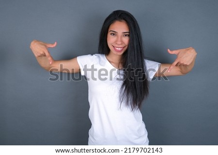Pick me! Confident, self-assured and charismatic young beautiful brunette woman wearing grey T-shirt over white wall promoting oneself as wanting role smiling broadly and pointing at body.