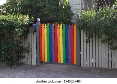 Picket fence in front of a weatherboard house. The gate is painted in colourful gay pride colours. Red; Life, Orange; Healing, Yellow; Sun, Green; Nature, Royal Blue; Harmony, Violet; Spirit.