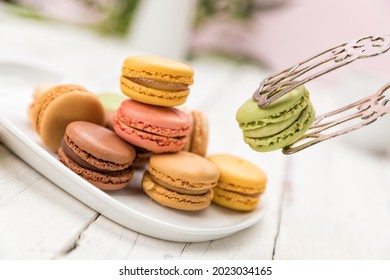 Picking A Pistachio Flavored Macaron With Pastry Tongs From A Selection Of Cookies On A Plate, Tilted View