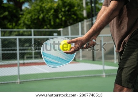 Pickleball player hand preparing to serve yellow ball, holding paddle on outdoor ourt during active fitness recreation gameplay training match exercise