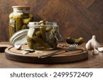 Pickled cucumbers in open jar, fork and spices on wooden table