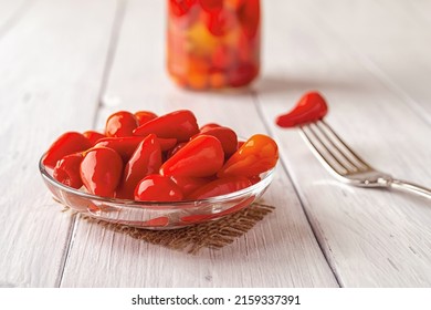 Pickled red hot peppers on a glass saucer over rustic wooden table. Marinated pods of small hot pepper close-up. Fermented vegetable snacks and condiments. Tasty pickles. Front view.